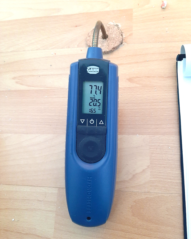 Zu sehen ist ein Hygrometer zu Messung der Bodenfeuchte, wie es mit dem Sensor in den Laminatboden gesteckt ist. Es zeigt eine Feuchte von 77,4 Prozent bei einer Temperatur von 20,5 Grad Celsius.